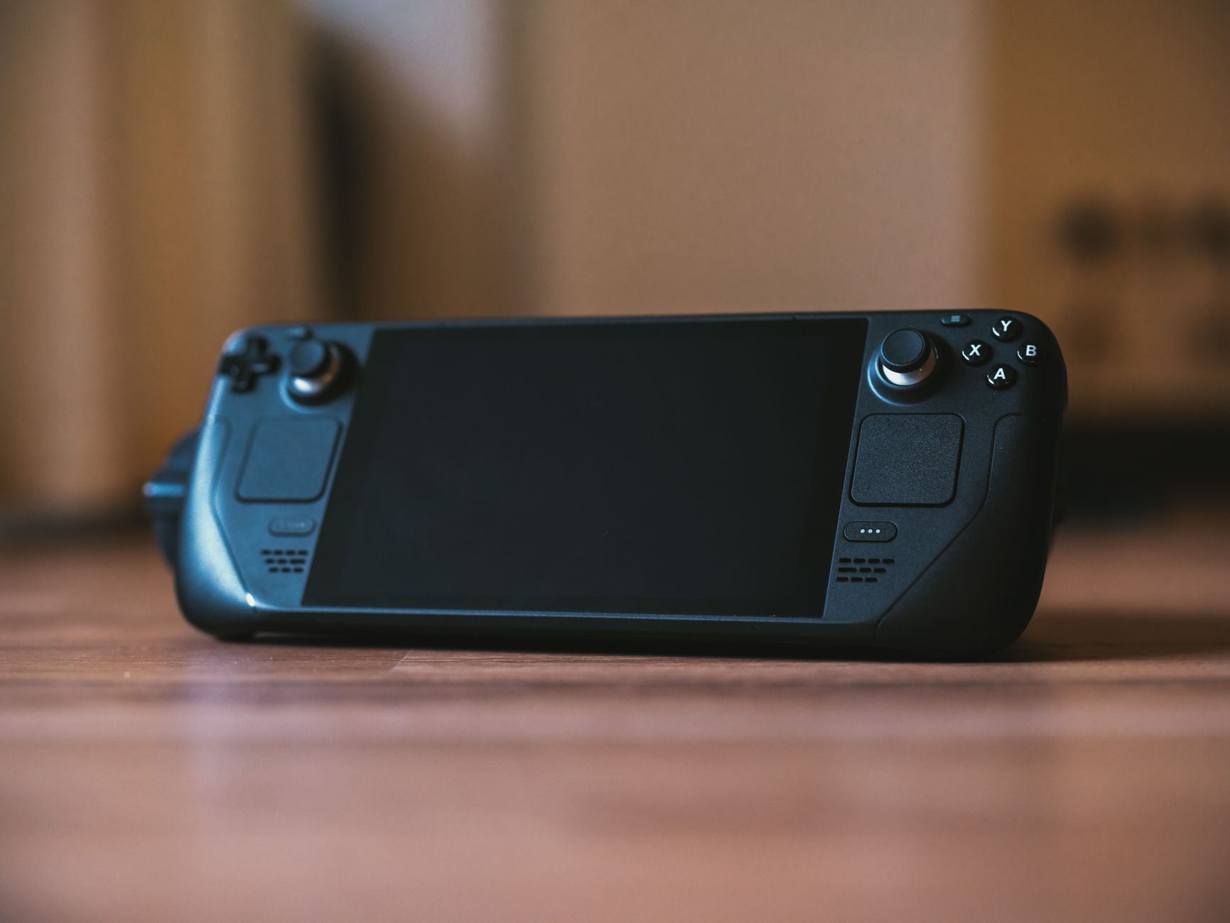 Console de jeu portable noire, écran éteint, posée sur une surface en bois devant un arrière-plan flou.