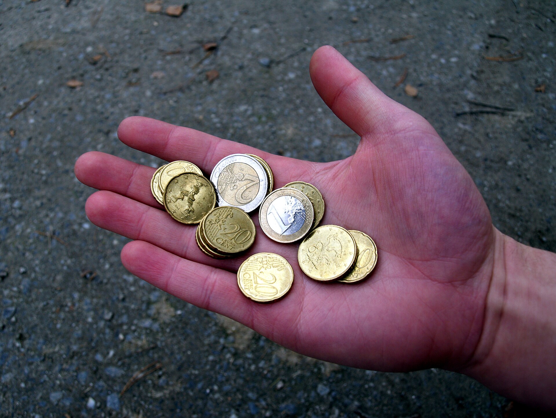 Main ouverte tenant une poignée de pièces en euros, dont des pièces de 2 euros, 1 euro et 20 centimes, au-dessus d'un sol de bitume.