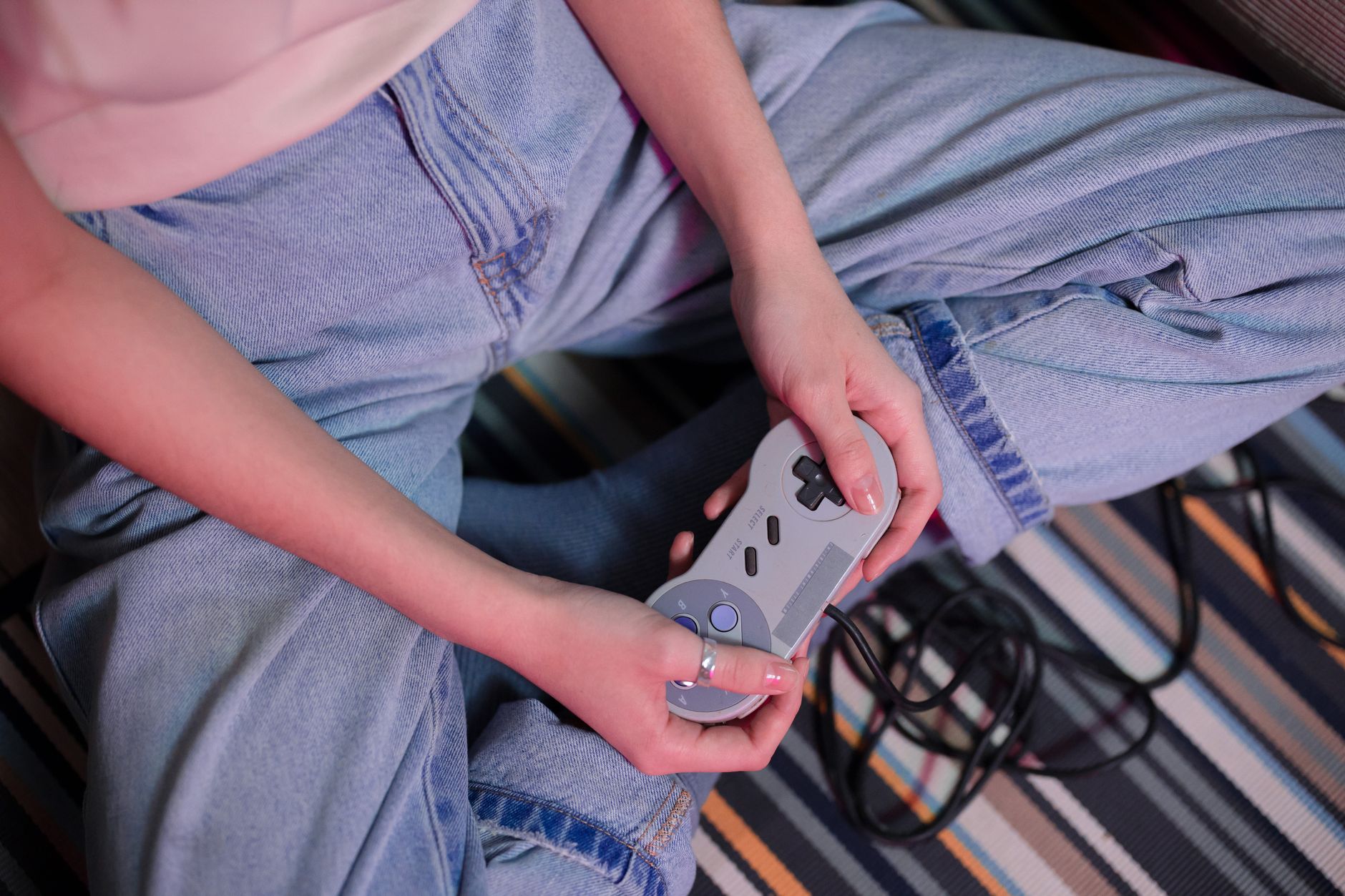 Personne en jean assise sur un tapis rayé, tenant une manette de jeu rétro grise dont le câble noir traîne au sol.