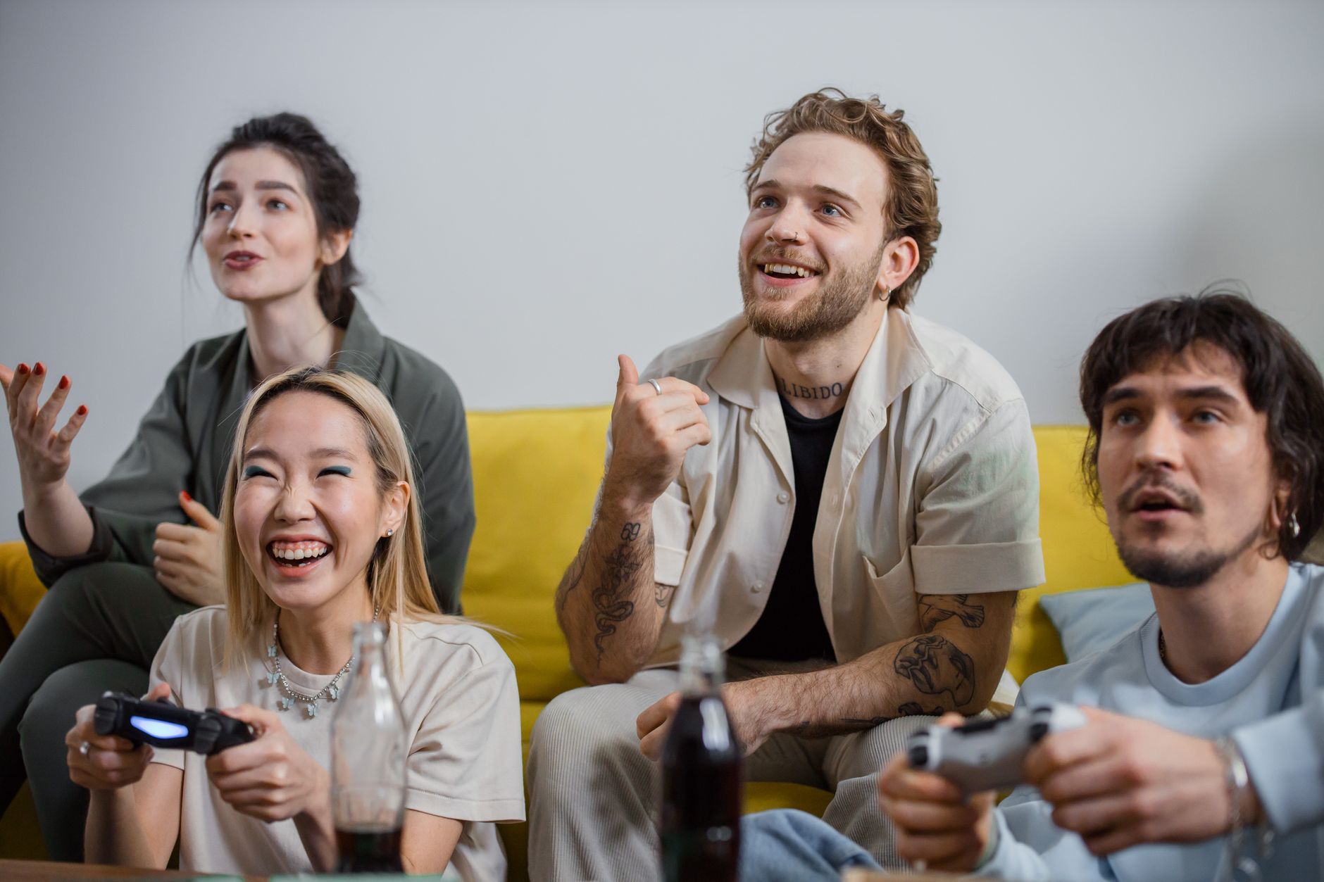 Quatre amis sur un canapé jaune jouent aux jeux vidéo avec des manettes, certains rient, des bouteilles de soda au premier plan.